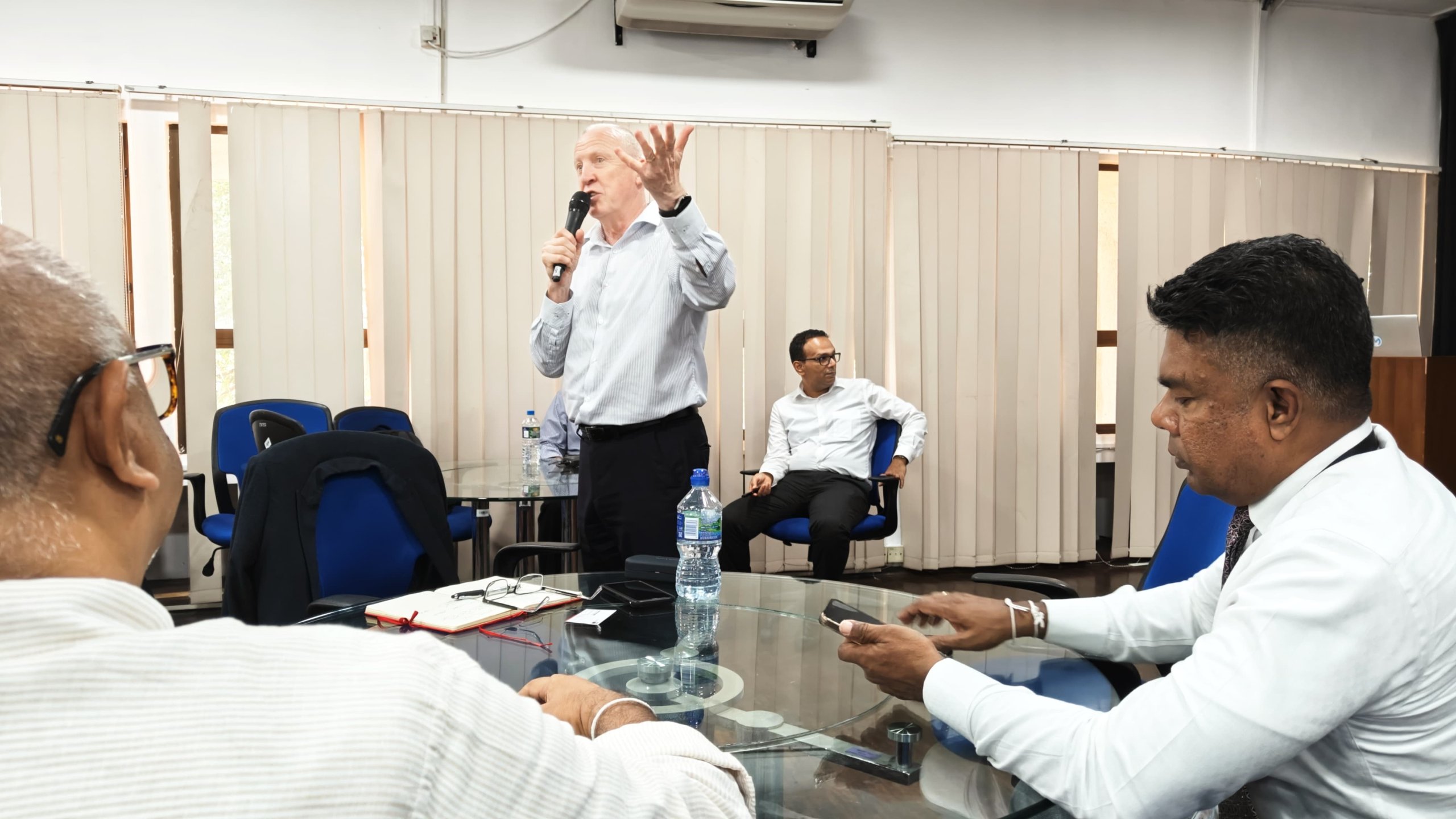 A man stands and speaks into a microphone in a meeting room, gesturing with his left hand. Three other men sit around a glass table with notebooks, a water bottle, and phones. Vertical blinds cover the windows behind them.