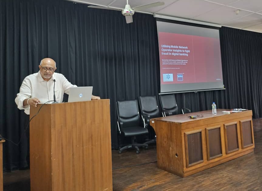 A man stands at a wooden podium with a laptop, delivering a presentation in a room with dark curtains. Behind him, chairs and a table are arranged. A projector displays a slide titled "Utilizing Mobile Network Operator Insights to fight Fraud in digital banking.