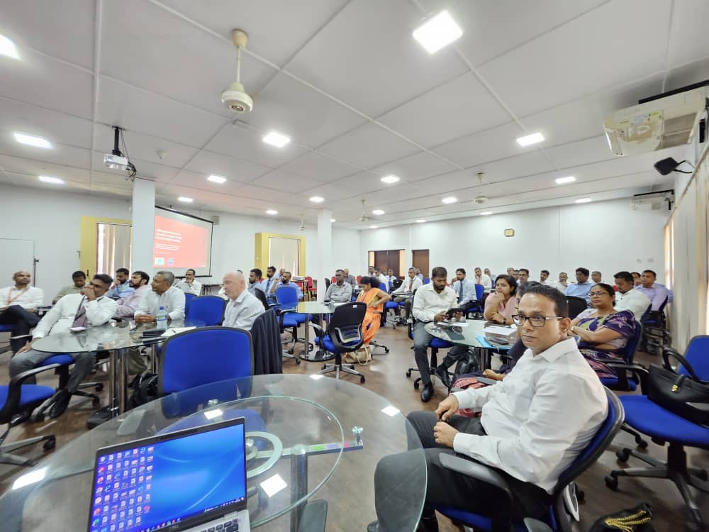 A group of people, mostly men in formal attire, sit at round glass tables in a bright conference room. Some look toward the camera, while others face a projector screen displaying a presentation. Laptops and bags are on the tables. The atmosphere is attentive.