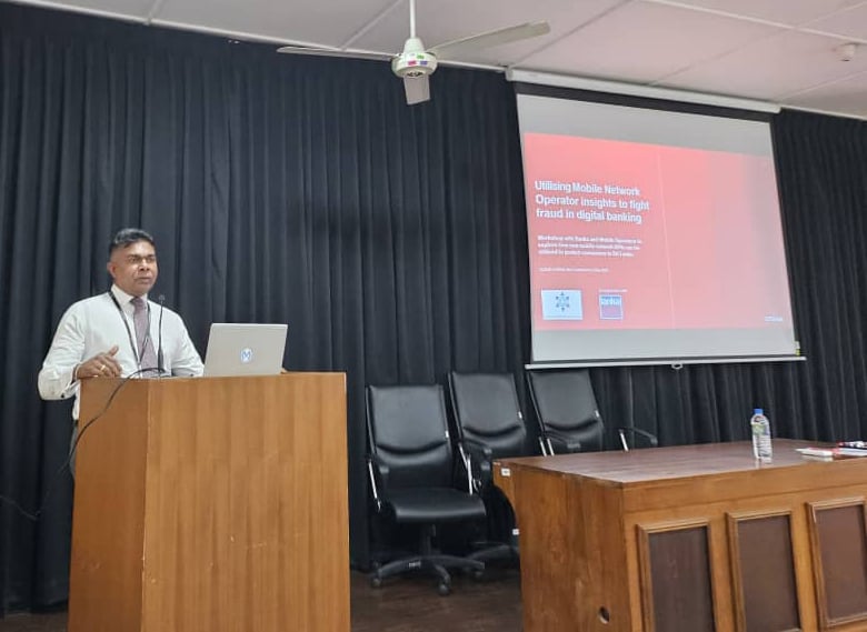 A man stands at a podium with a laptop, presenting in a room with black curtains. A slide on the projector screen reads “Utilizing Mobile Network Operator Insights to fight fraud in digital banking.” Two empty chairs and a wooden table are beside him.