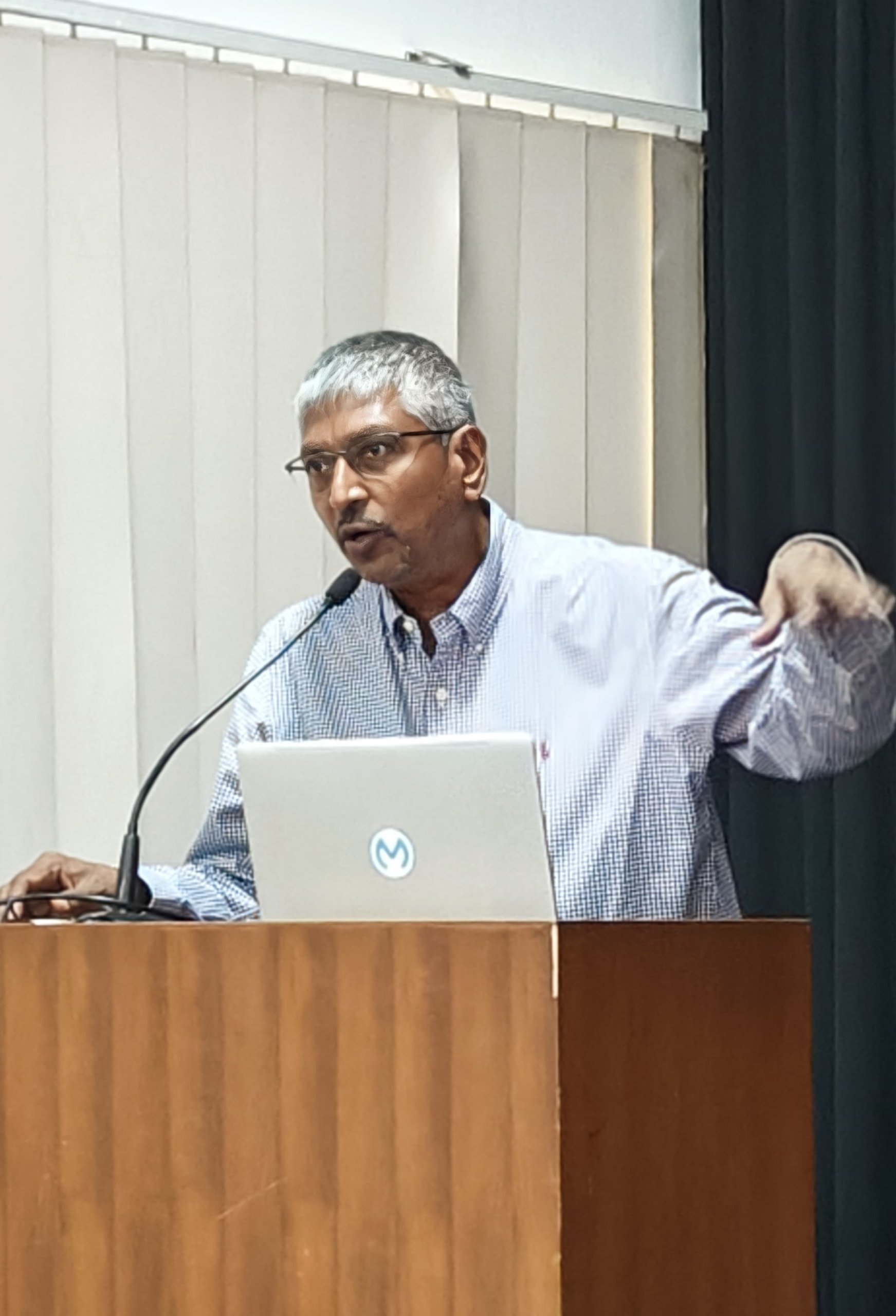 A man with short gray hair and glasses speaks passionately at a podium, gesturing with his right hand. He is wearing a light checked shirt and stands behind a laptop. Vertical blinds and a dark curtain are visible in the background.
