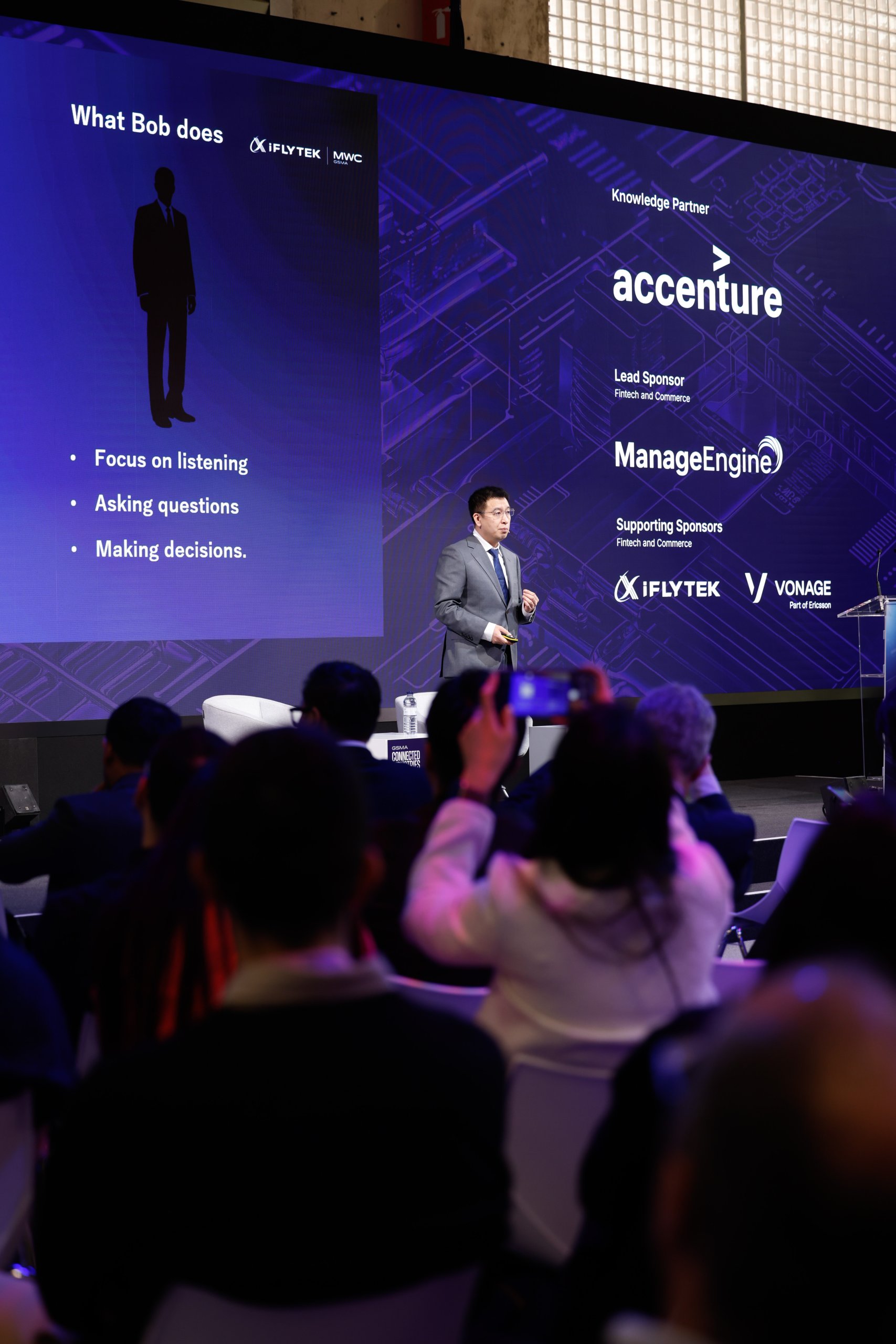Man giving a presentation on stage; screen behind lists tasks and event sponsors; audience watching.