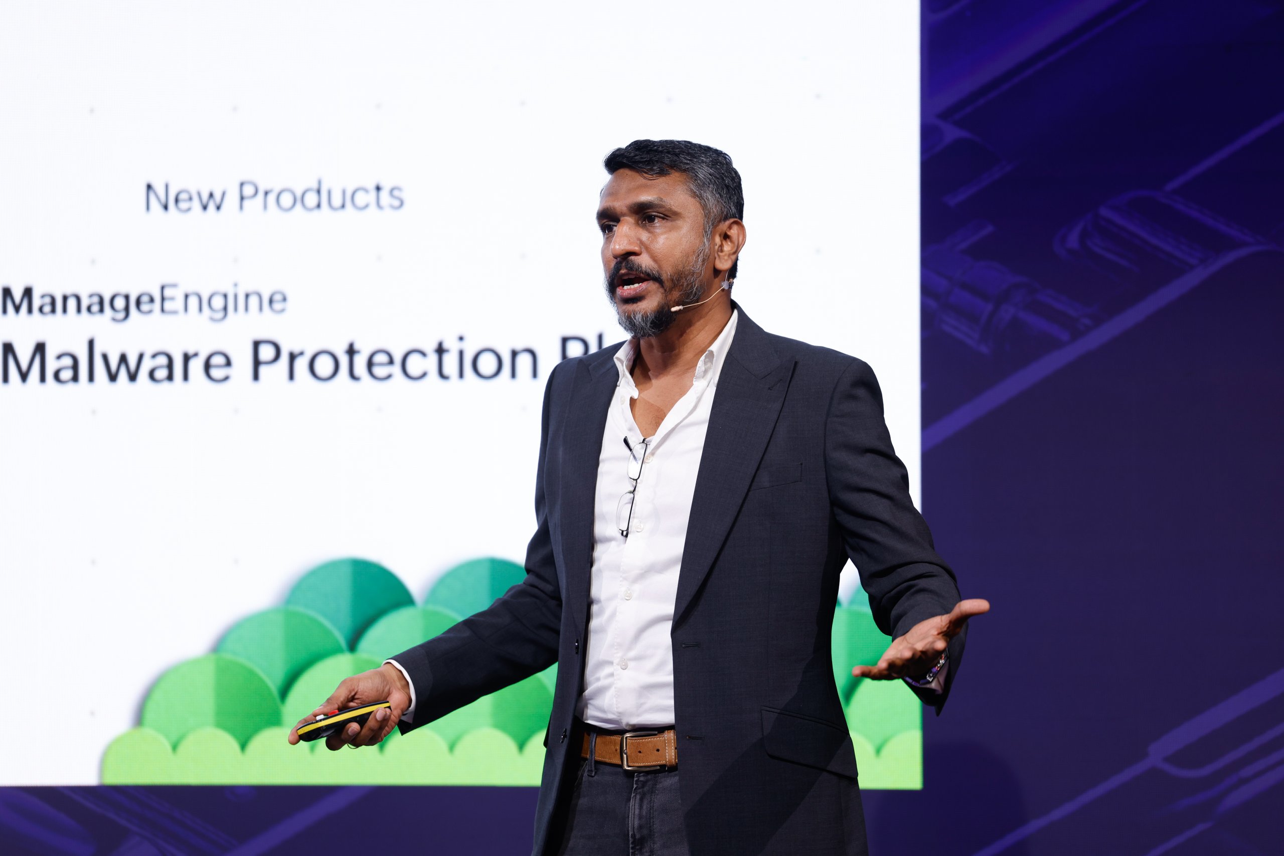 A man in a suit speaks on stage with a "Malware Protection" presentation shown behind him.