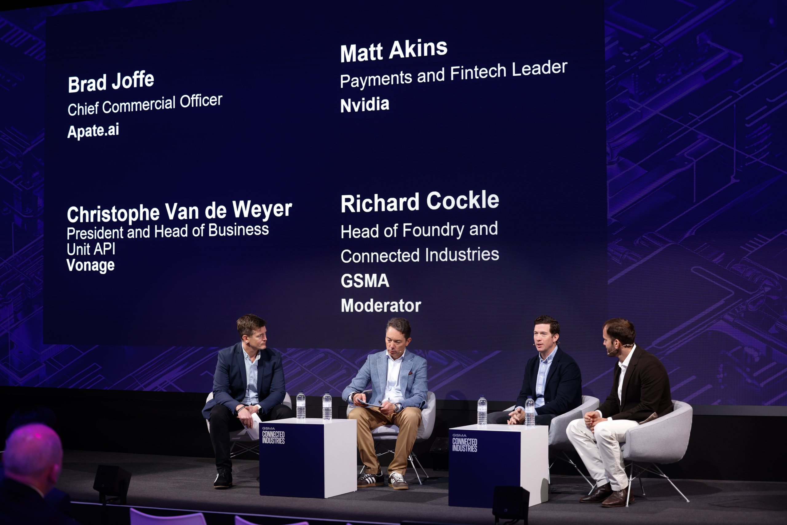 Four men in suits sit on a stage panel discussing technology, with their names and titles displayed behind them.