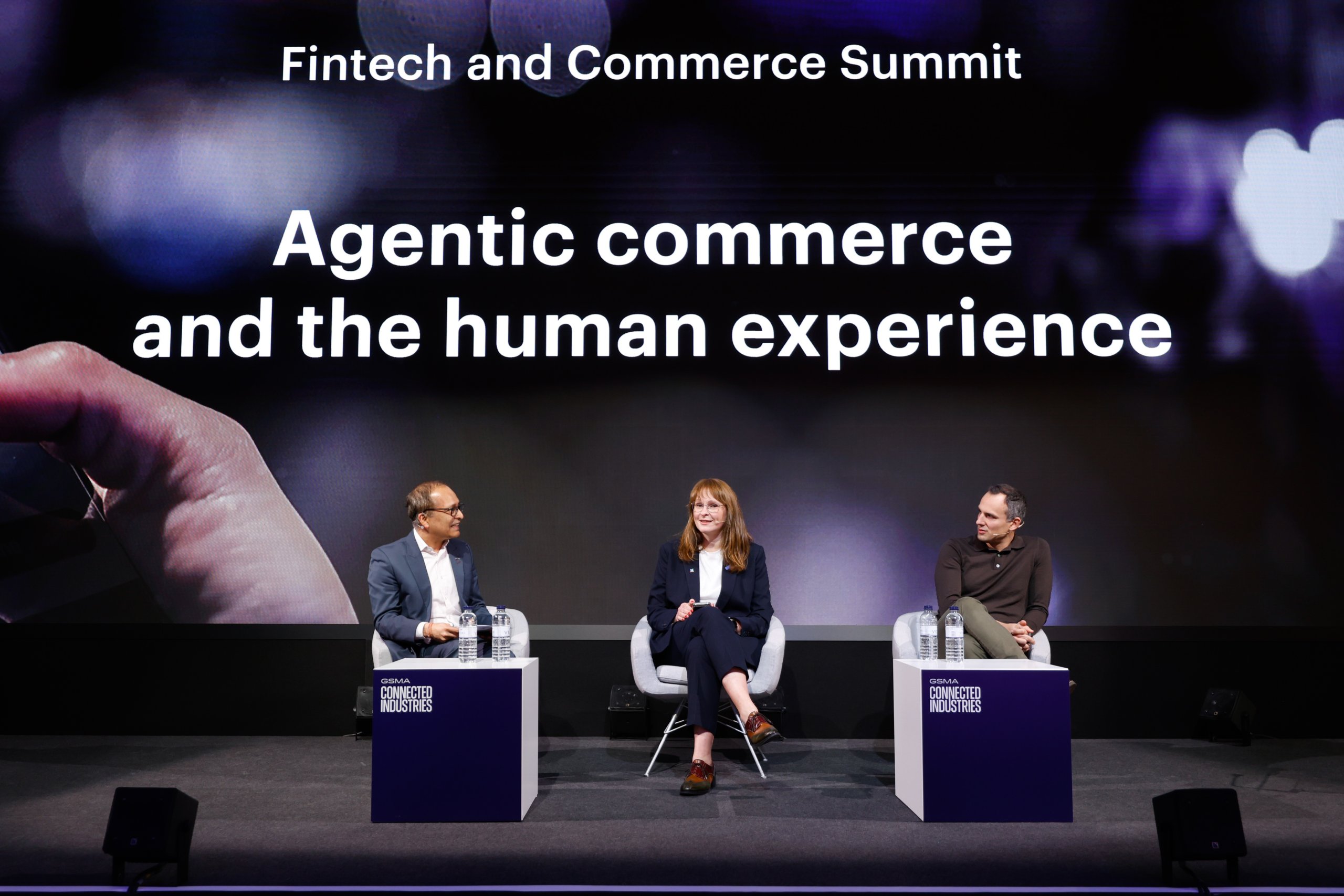 Three people sit on stage at the Fintech and Commerce Summit with a large display reading, “Agentic commerce and the human experience.”.