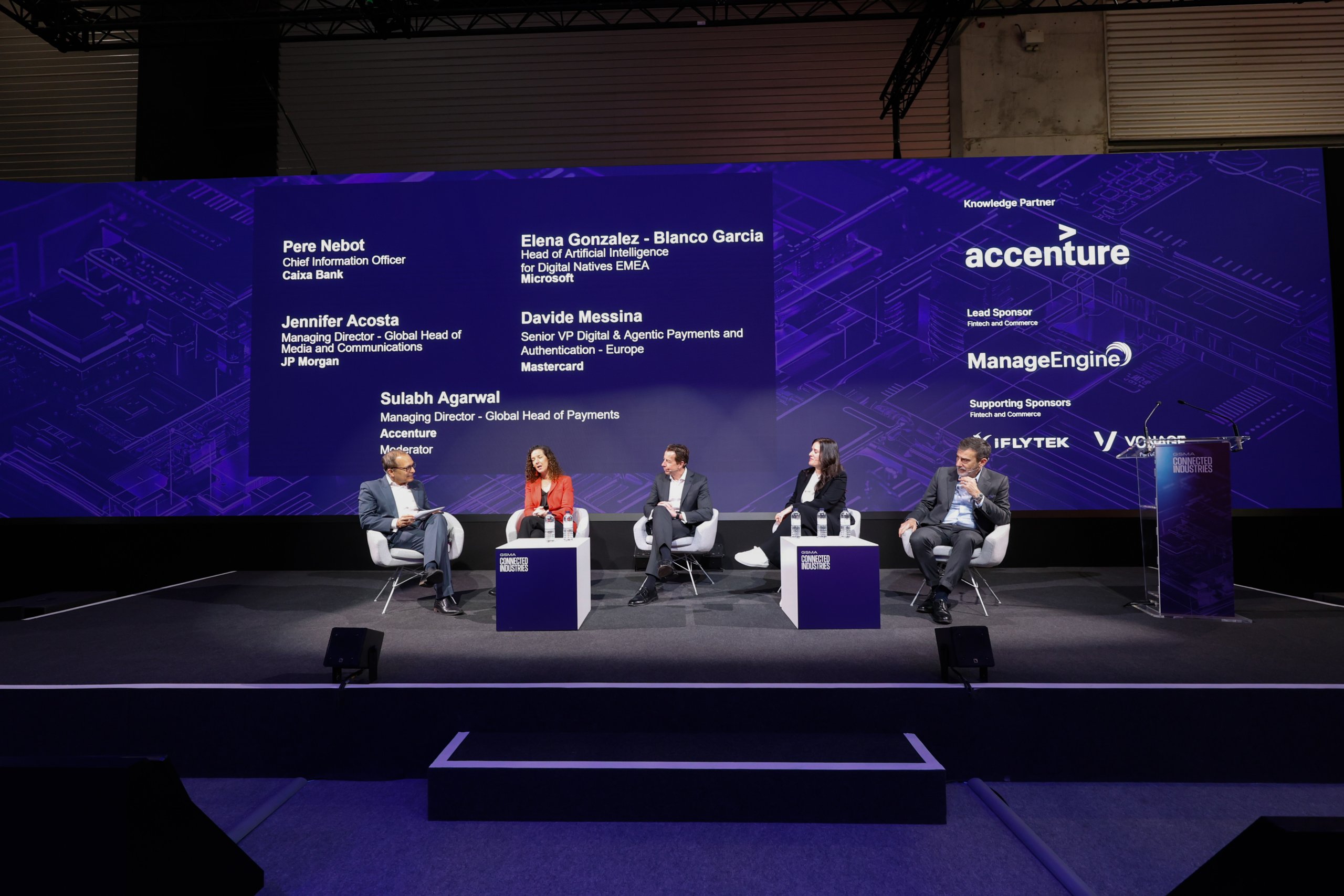 Five panelists sit on stage in front of a screen displaying their names and companies at a business conference.