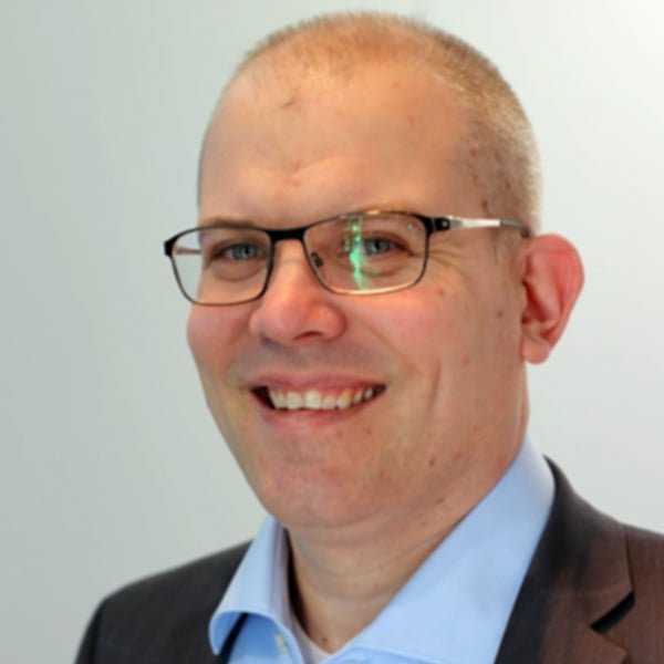 A smiling person with short, light hair and glasses wearing a light blue shirt and a dark suit jacket, photographed against a plain, light background. The person is facing forward, and the lighting is even.