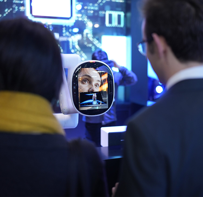 Two people are standing in front of a futuristic display showing a screen with a human face on it. The background is filled with blue lights and electronic elements, creating a high-tech atmosphere. One person in the background is adjusting equipment.