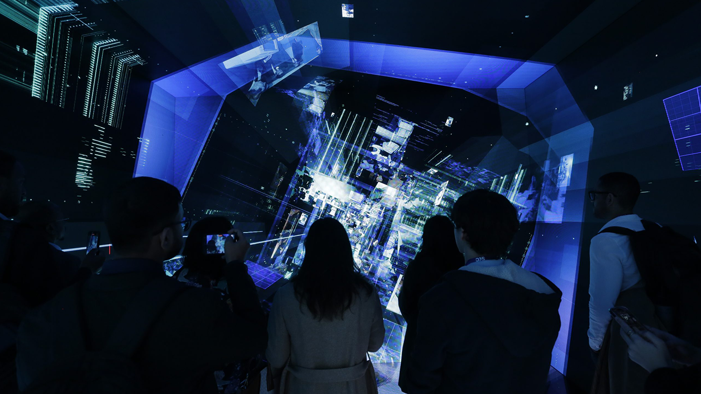 Several people are standing in a dark room, observing a large, illuminated digital display showing a complex network of interconnected lines and shapes. Some people are holding up their phones, possibly taking pictures or recording videos.