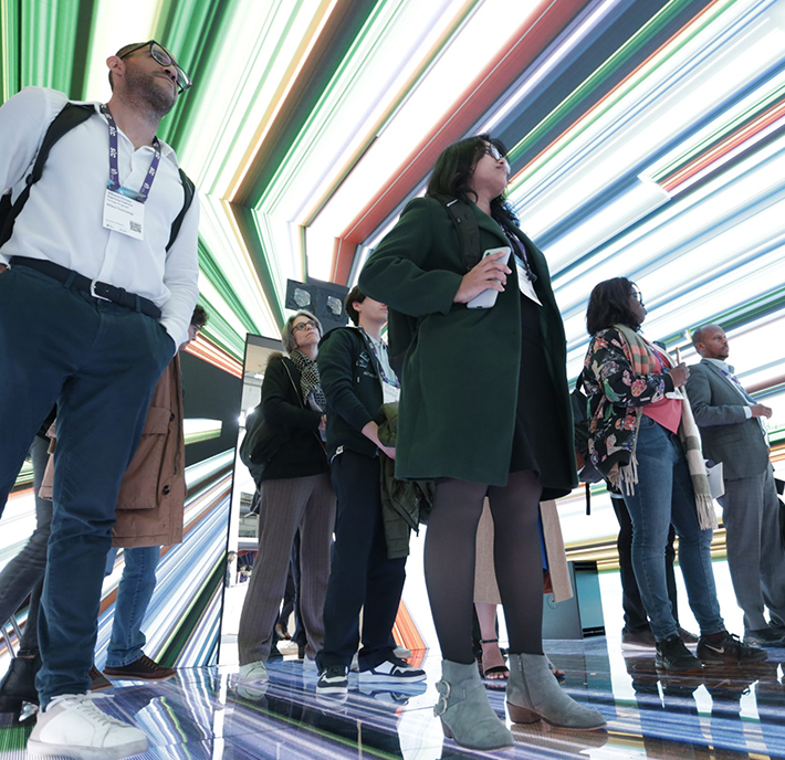 A group of people standing on a reflective floor, looking up at an installation of colorful, striped lights on the ceiling. Some individuals wear lanyards, indicating they might be at an event or presentation. The atmosphere appears modern and dynamic.