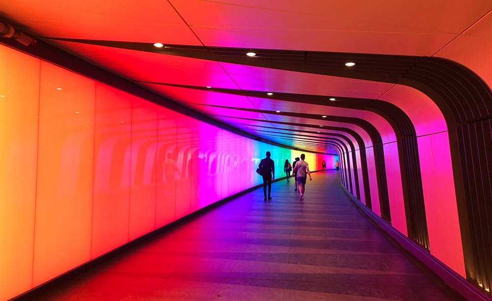 A curved hallway lit with vibrant, gradient rainbow colors along the left wall. Silhouetted people walk through the tunnel, where smooth panels and ceiling lights evoke a futuristic, colorful atmosphere—like stepping into an AI in mobile-inspired world.
