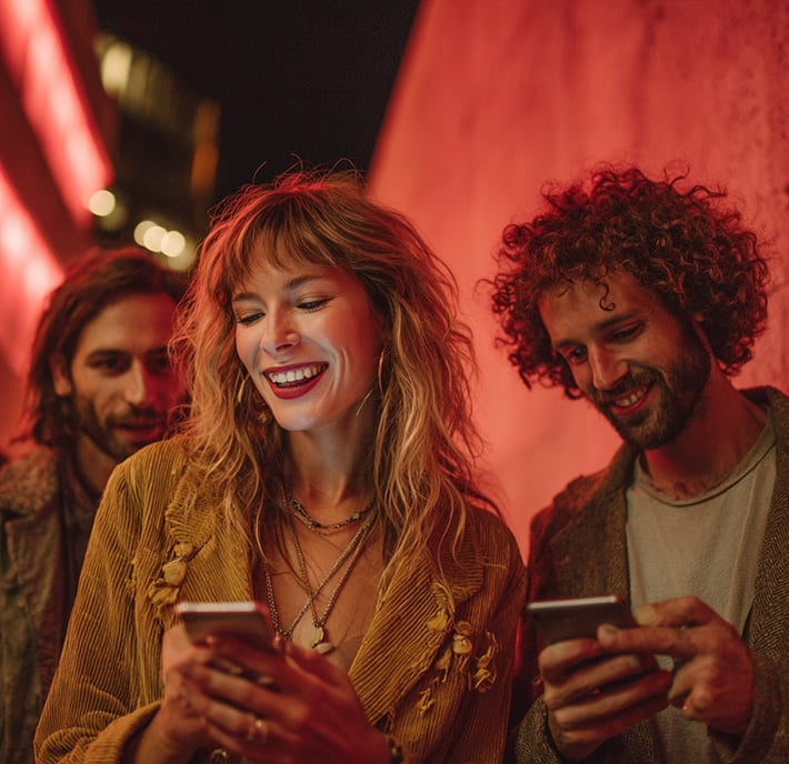 Three young adults stand together at night next to a red-lit wall, all smiling and looking at their phones. The woman in the center has wavy blonde hair and wears layered necklaces and a tan jacket; the two men beside her have curly and wavy hair.