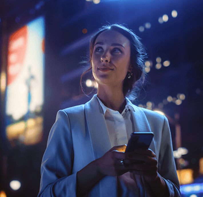 A woman in a light blazer stands outdoors at night, smiling softly while holding a smartphone. Neon lights and blurred billboards illuminate the cityscape behind her, creating a vibrant, urban atmosphere.