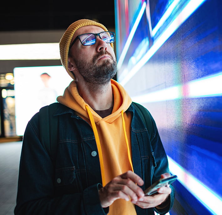 A bearded man in glasses, a yellow beanie, and a yellow hoodie under a dark jacket looks up thoughtfully at a bright digital screen. He holds a smartphone and stands indoors, with colorful lights reflecting on his face.