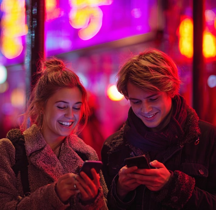 Two young people stand outside at night, smiling and looking at their smartphones. Neon pink and purple signs glow in the background, casting colorful light on their faces. Both wear warm jackets and appear happy and engaged with their devices.