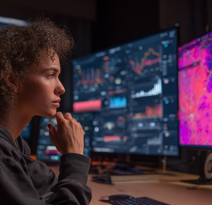 A person with curly hair sits at a desk, thoughtfully looking at several large monitors displaying complex graphs, charts, and colorful data visualizations in a dark room, suggesting analysis or monitoring of advanced digital information.