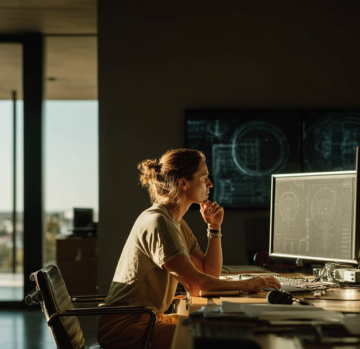 A woman sits at a desk in a modern office, intently looking at technical diagrams on a large computer monitor. Sunlight streams in as two screens display blueprints, hinting at engineering or architecture work possibly enhanced by AI in mobile technology.
