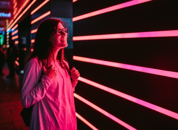 A person wearing glasses and a light long-sleeve shirt stands smiling with their backpack straps in hand, illuminated by pink and red neon horizontal lights on a dark urban wall, capturing the vibrant scene with mobile AI technology.