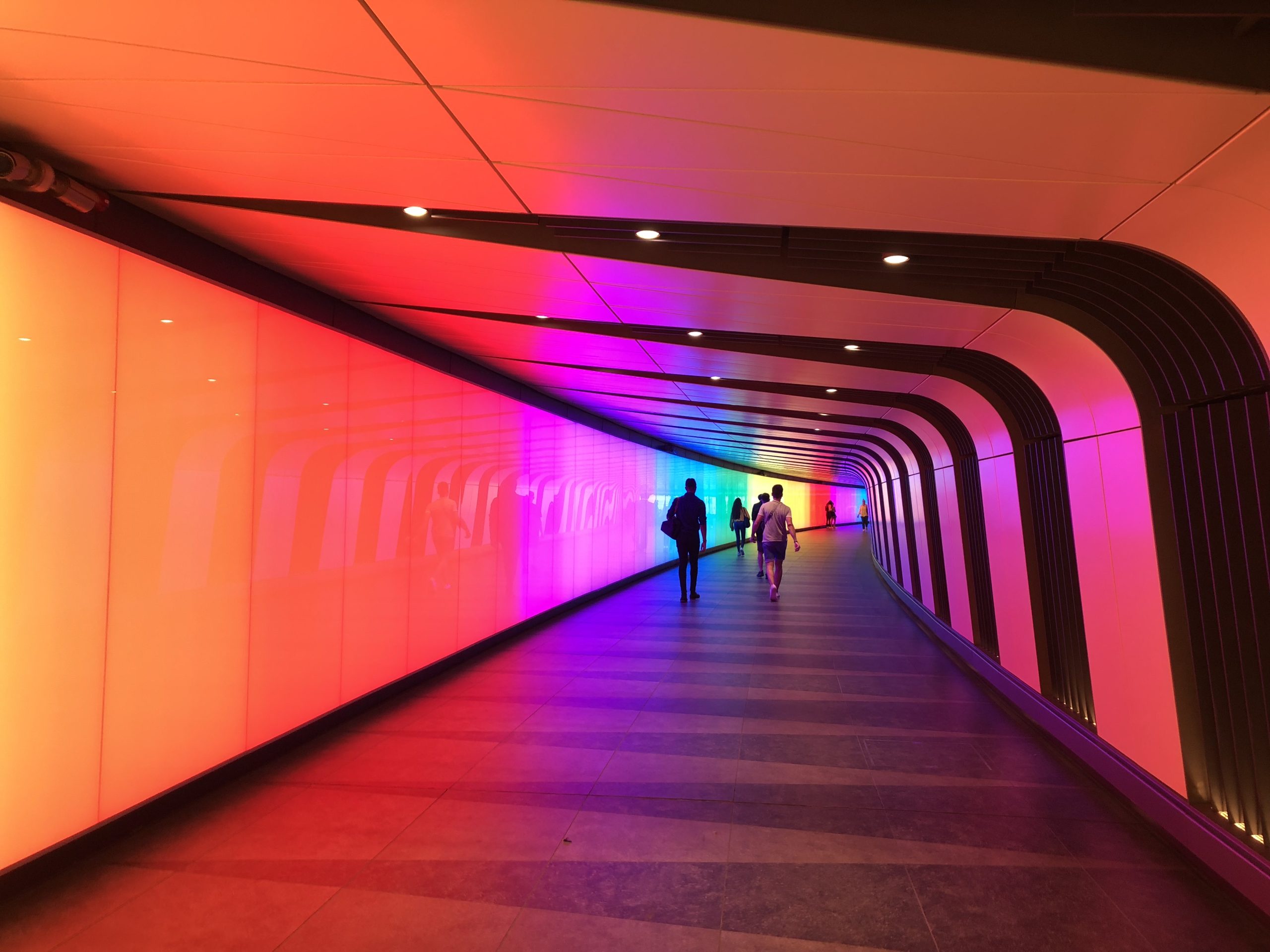 A modern indoor tunnel with a curved wall illuminated by vibrant gradient lights in orange, pink, purple, and blue evokes the feeling of LLM Benchmarks. Four people walk through the corridor’s clean tile floor and dark ceiling beams, creating a colorful, futuristic vibe.