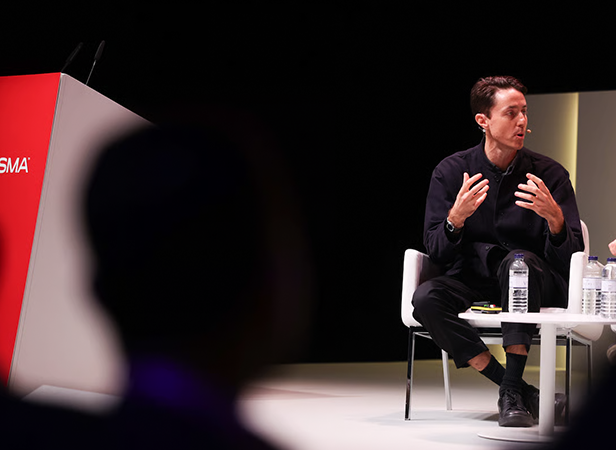 A person sits on stage speaking and gesturing with hands during a panel discussion about AI agents, with water bottles nearby.