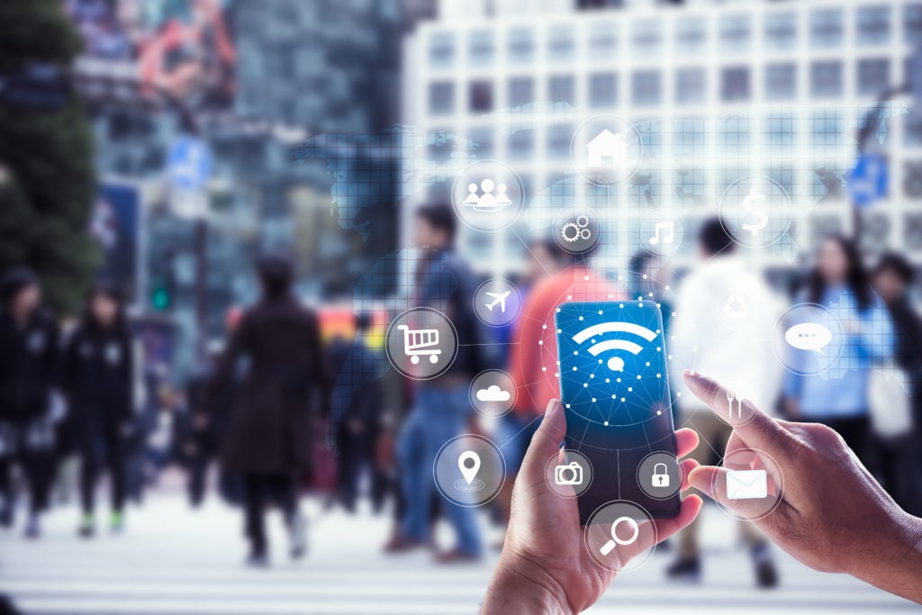 A person holds a smartphone with various digital icons, such as Wi-Fi, shopping cart, and music, floating around it. In the background, a busy urban street scene with blurry pedestrians walking, suggesting a connection between mobile technology and everyday life.
