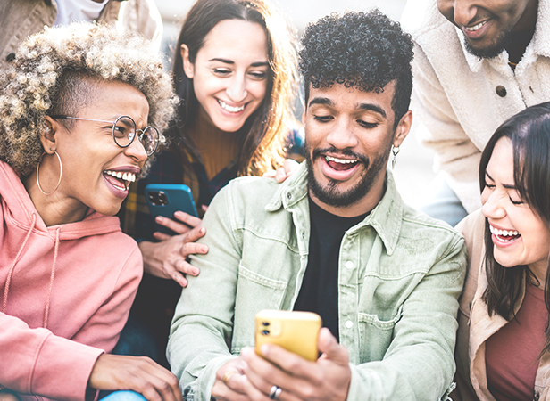 A diverse group of five young adults laughing and looking at a smartphone together outdoors.