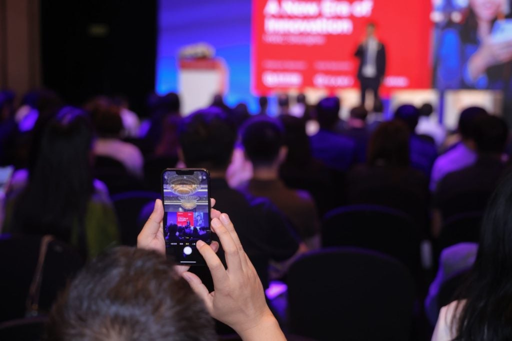A person in the foreground is taking a photo with a smartphone at a conference. The audience is seated and facing a brightly lit stage where a speaker stands. A large screen displays text about innovation in the background. The setting is a dimly lit auditorium.