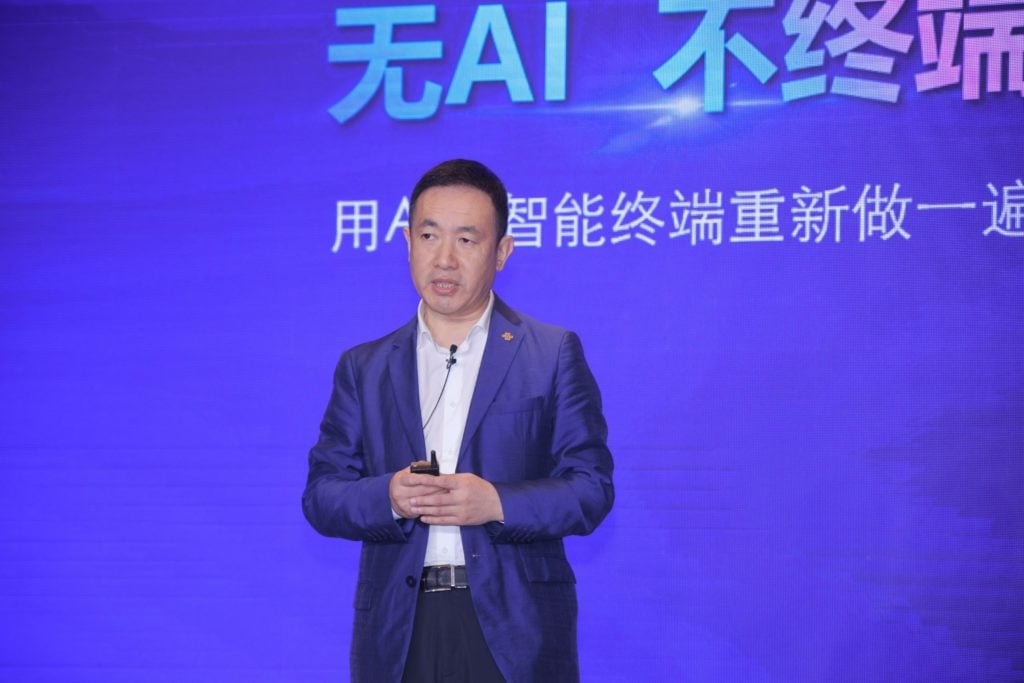 A man in a blue suit stands on a stage holding a microphone. He is presenting against a blue and purple background with Chinese text. The text is partially visible, and the lighting highlights the speaker while casting soft shadows.