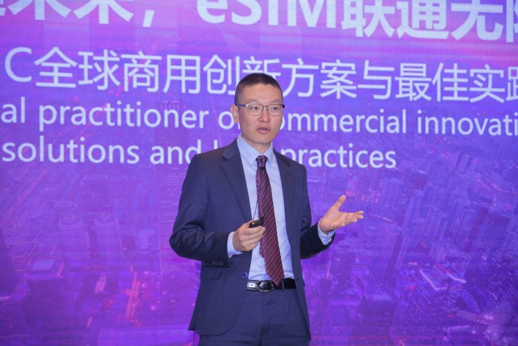 A man in a suit and tie speaks into a microphone, holding a presentation clicker in one hand. He stands in front of a purple-lit background displaying Chinese text and English phrases about global commercial innovation and best practices.