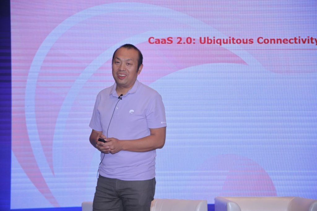 A man in a light purple polo shirt stands on stage, holding a remote, with the screen behind him displaying the text "CaaS 2.0: Ubiquitous Connectivity." Two empty white chairs are partially visible at the bottom of the image.