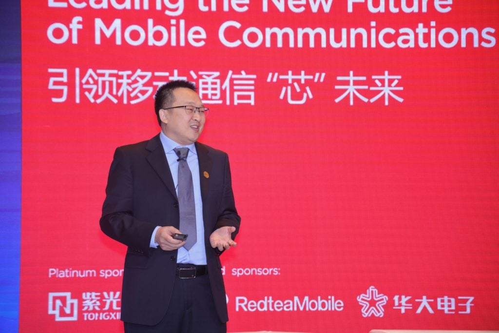 A man in a suit stands on stage delivering a presentation. Behind him, a red screen displays text in English and Chinese, including phrases about leading mobile communications. Logos of sponsors are visible at the bottom. The setting suggests a formal conference or event.