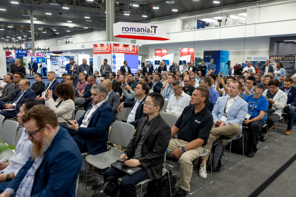 A large group of people sits in rows at a tech conference, focusing on a presentation. Attendees are diverse in age and business attire. In the background are booths and banners, including one for "romaniaIT." The room is well-lit, with several people standing near the exhibits.