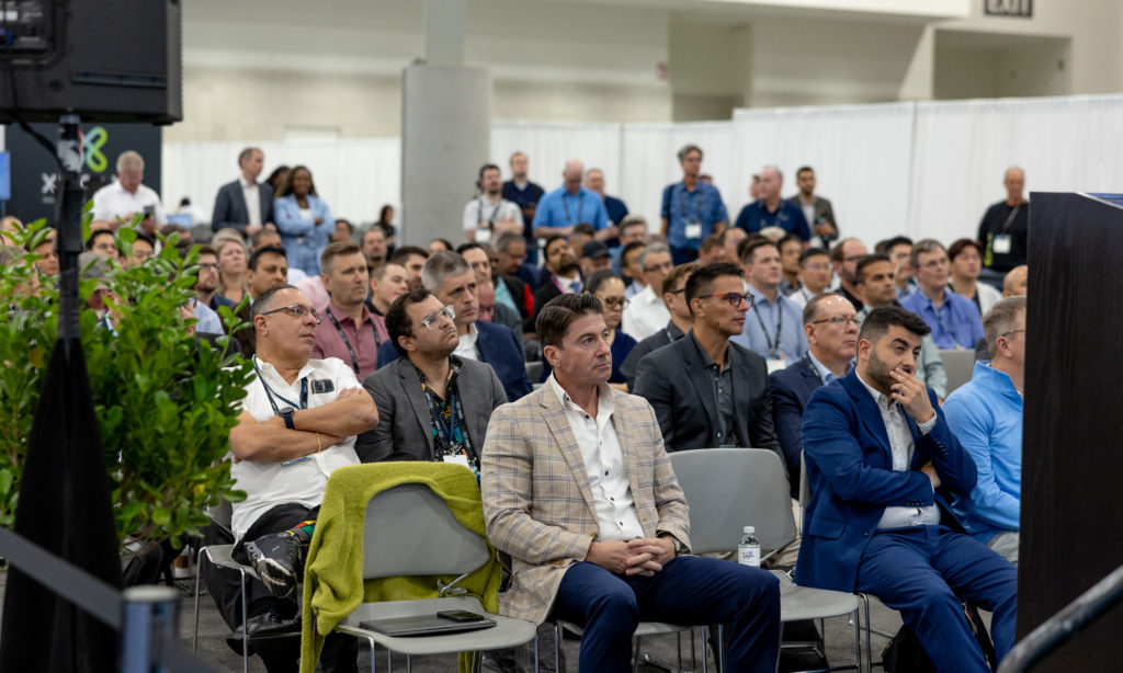 A large group of people is seated attentively in a conference room, facing a speaker off-camera. Most are middle-aged men in business attire, with some wearing glasses. A few women are present. Green plants are in the foreground. White curtains and exhibition panels are in the background.