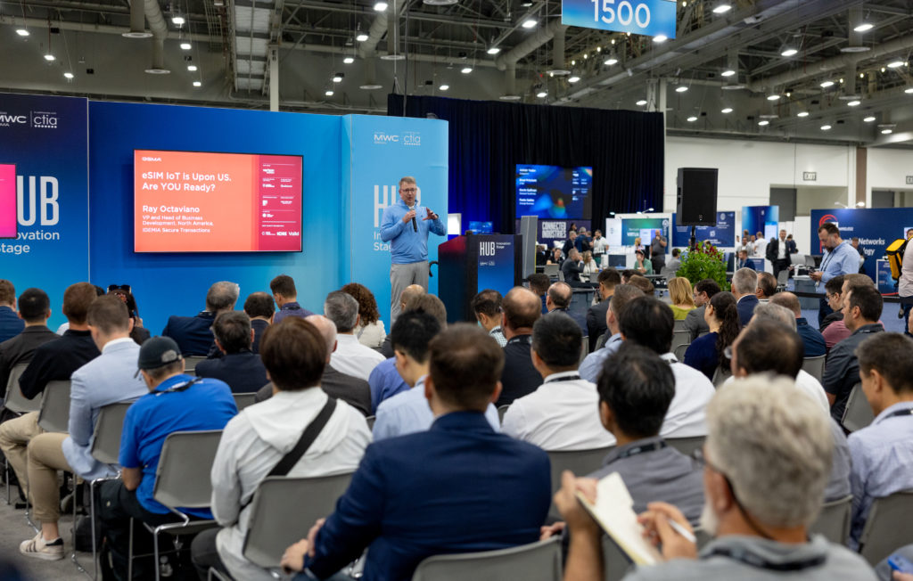 A man speaks on stage at a tech conference in a large exhibition hall. Attendees sit and listen, some taking notes. A screen displays a presentation titled "eSIM IoT is Upon Us, Are You Ready?" The venue is filled with booths and digital displays, with a bustling audience.