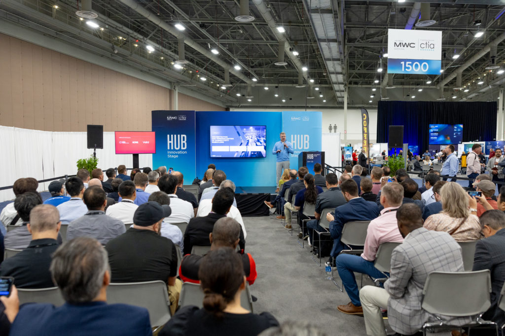 A speaker is presenting on stage at a tech conference with a large audience engaged. The digital display highlights eSIM technology, while "HUB" and "MWC24 Las Vegas" banners are visible. Attendees use laptops and phones in the spacious venue, setting the tone for a cutting-edge discussion.