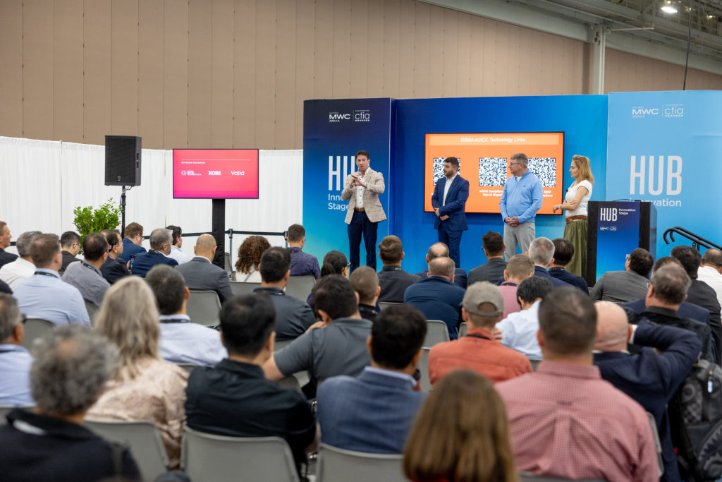 A speaker addresses an audience at a conference in a large hall. Behind him, a screen displays two QR codes and text about eSIM technology. Three other people stand nearby. Attendees sit closely, observing the presentation. The backdrop includes logos and the word "HUB" in large letters, hinting at MWC24 Las Vegas.