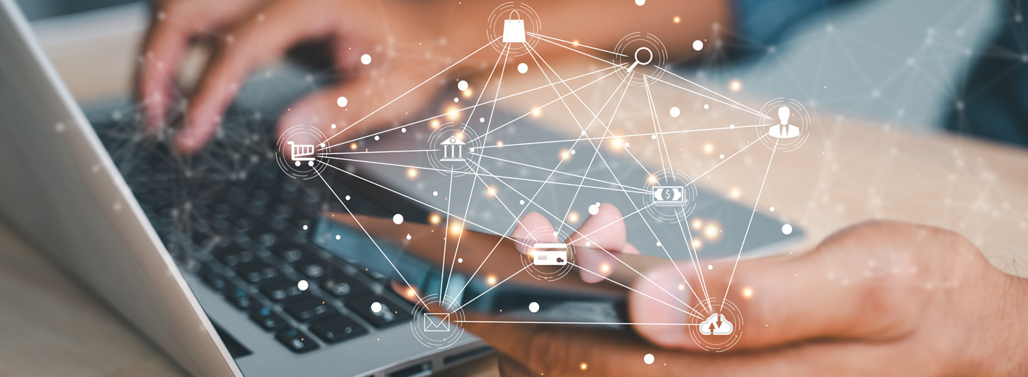 A person types on a laptop while holding a smartphone. Overlayed icons connected by lines include a shopping cart, bank, smartphone, card, envelope, and person, symbolizing digital connectivity and online transactions. A blurred background enhances the focus on the devices and symbols.