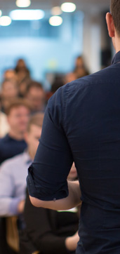 A person in a navy shirt is speaking to a seated audience in a conference room. The audience is slightly blurred, with some individuals visible. The room has bright overhead lighting, creating a professional and focused atmosphere. The speaker's back and arm are prominent in the foreground.