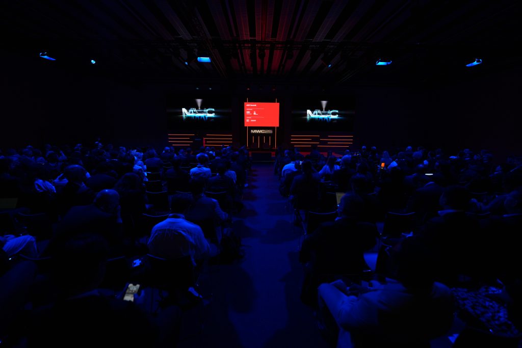 A room full of people sat in front of a stage with two big screen in front of them playing a video