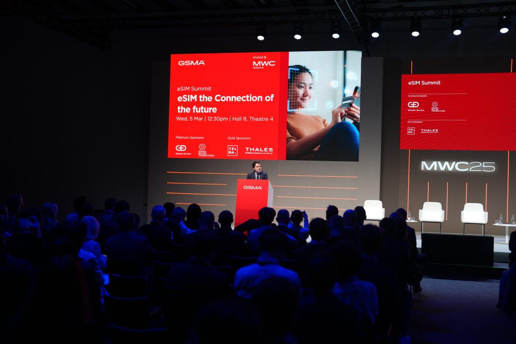 A man on stage behind a podium with a mic and a big screen behind him with a banner of a woman holding a mobile phone in her hand in a room full of people