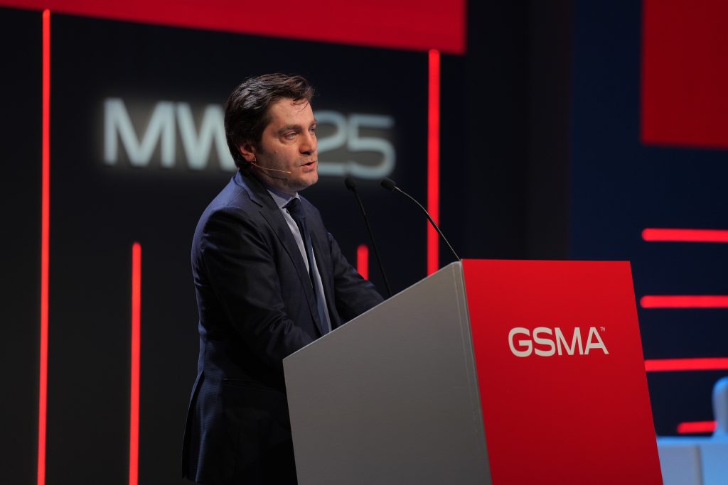 A man on stage standing behind a podium with a mic in front of him. On the podium is a red banner with GSMA written on it