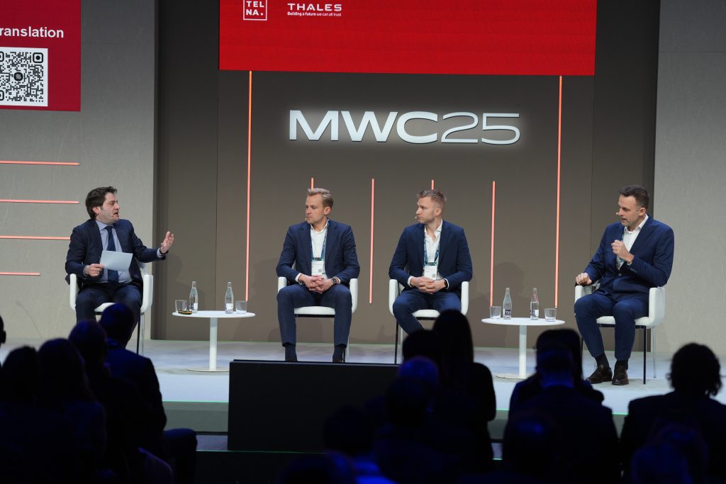 Four men on stage sitting down in front a audience of people. There are two small table on the stage with bottled water