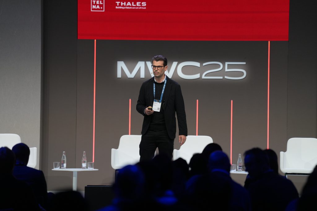 A man on stage holding a device in his hand and talking to a room full of people