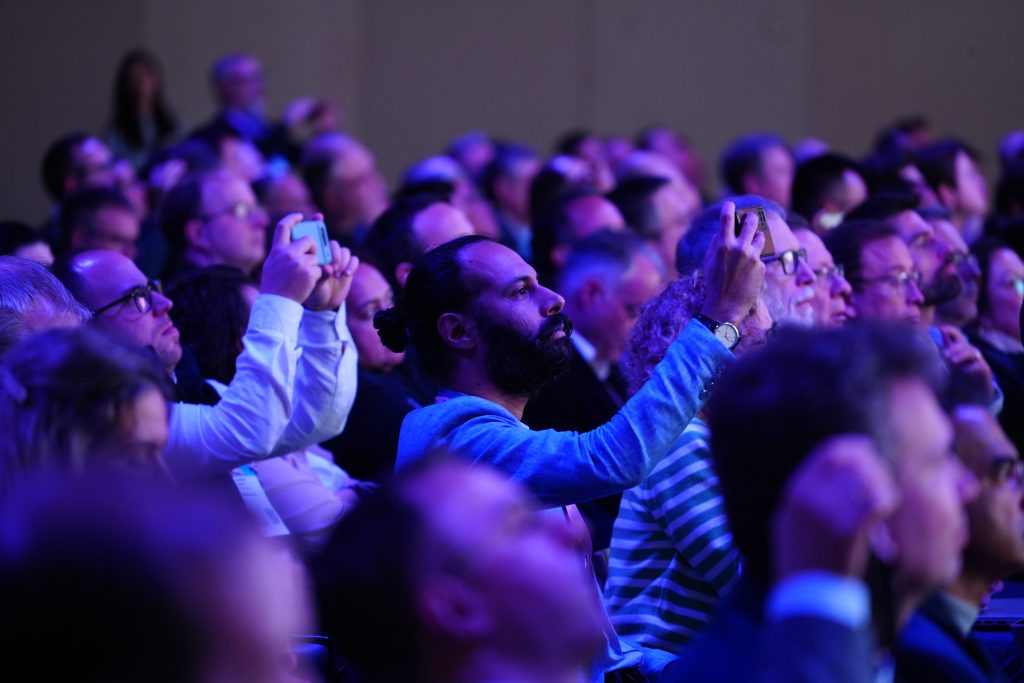 Audience members at an event, some holding up mobiles to take photos, under blue and purple lighting.
