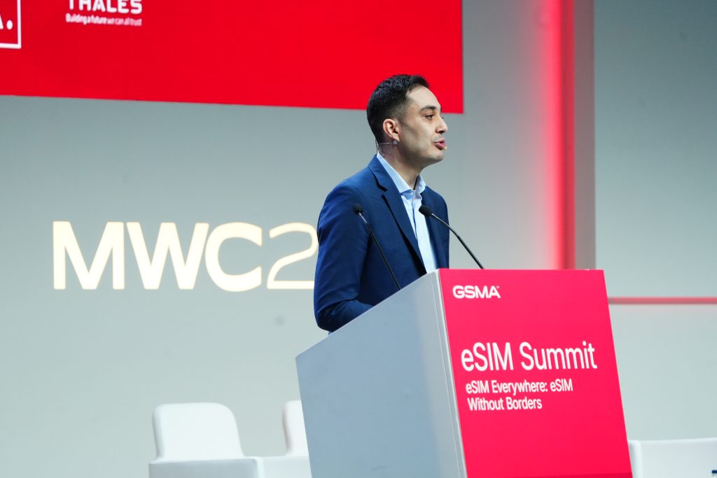 A man in a blue suit speaks at a lectern labelled "eSIM Summit" during the MWC event.