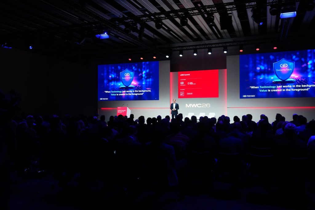 A speaker presents on stage at a tech conference, with slides and a large audience in a dark room.