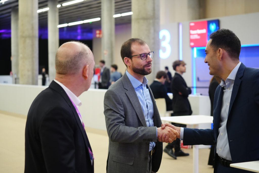 Three men in business attire shaking hands and talking at a modern indoor eSIM on-demand event or conference.