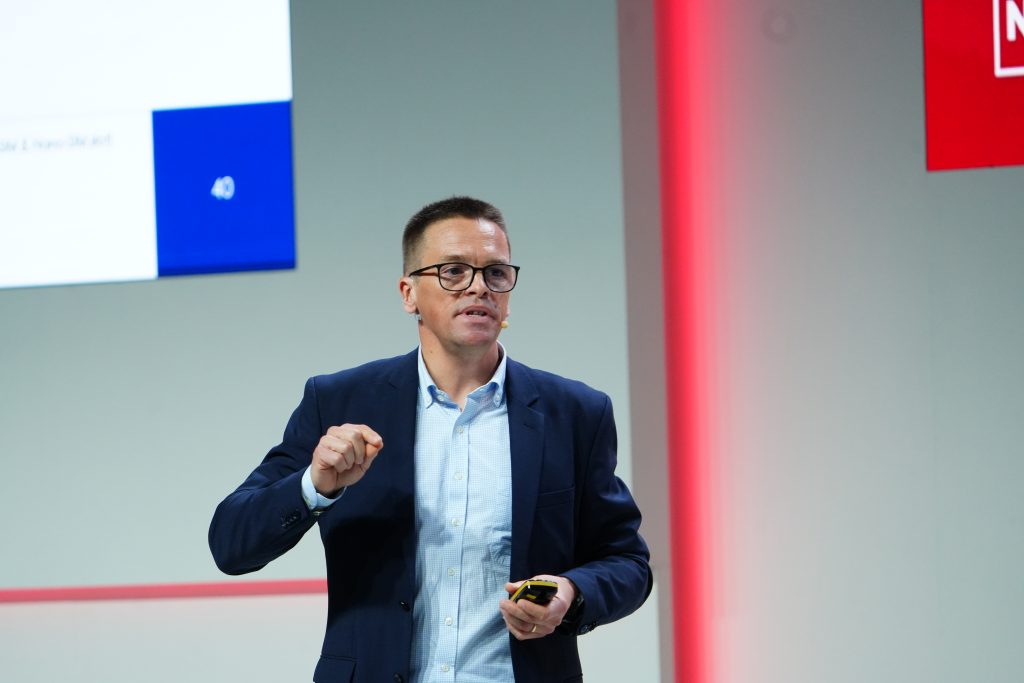 Man in a blue suit giving a presentation on stage, holding a remote and speaking.