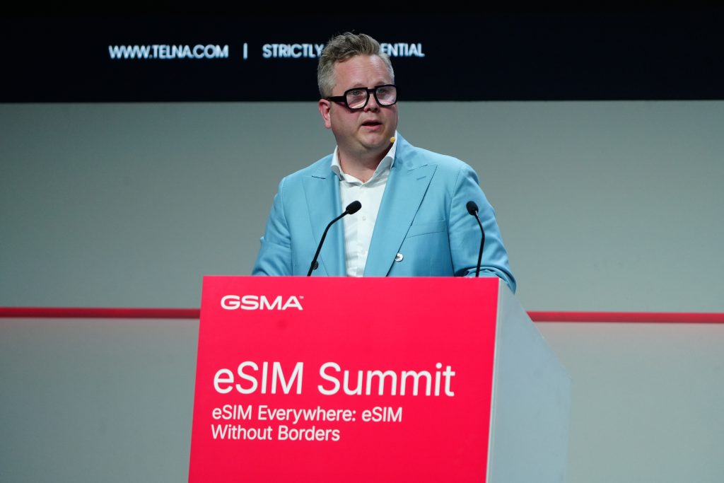 A man in a blue suit speaks at a lectern during the GSMA eSIM Summit, discussing innovations like SGP.32.