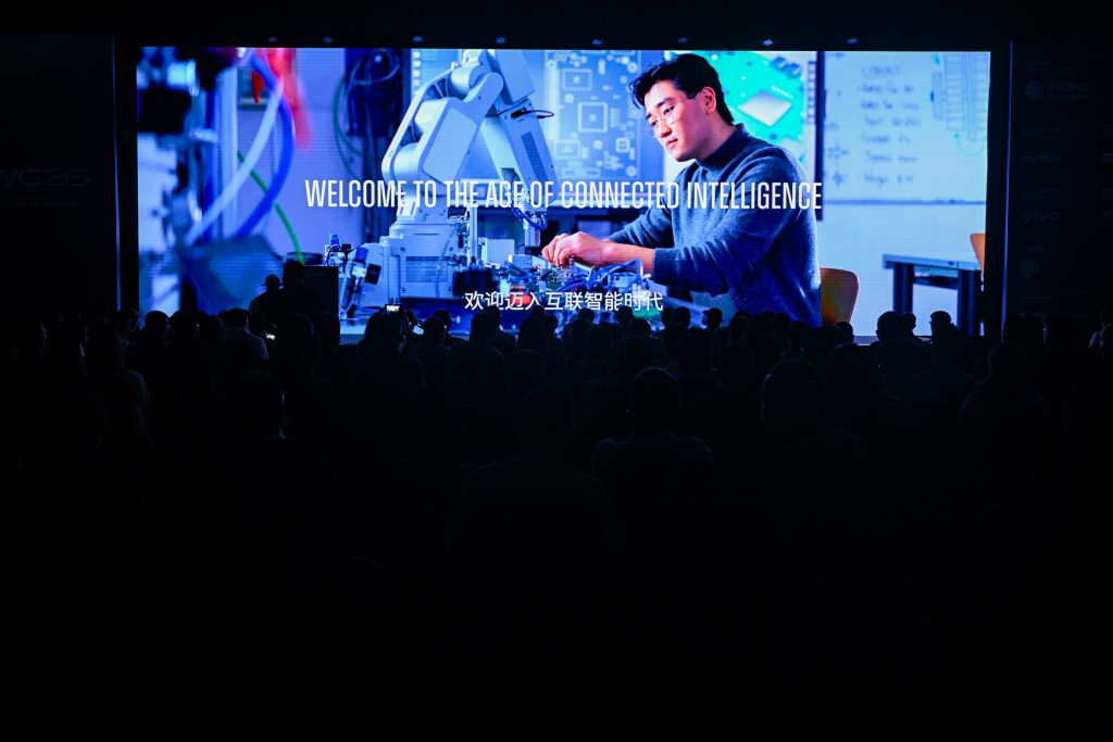 A dark auditorium audience watches a large screen showing a man working with robotics. The text “WELCOME TO THE AGE OF CONNECTED INTELLIGENCE” appears above Chinese subtitles. The background is brightly lit, contrasting with the silhouettes.